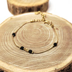 Bracelet doré en acier inoxydable avec perles noires facettées et perles dorées posé sur fond bois, bijou raffiné et moderne Clyvia Bijoux.