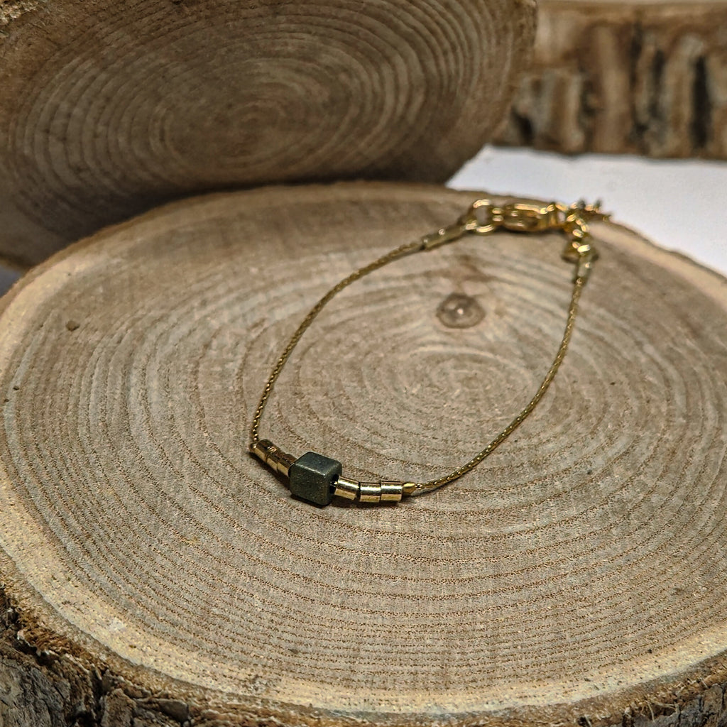 Bracelet fin doré avec perle irisée aux reflets bleu-vert posé sur bois, bijou raffiné et lumineux Clyvia Bijoux.