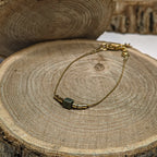 Bracelet fin doré avec perle irisée aux reflets bleu-vert posé sur bois, bijou raffiné et lumineux Clyvia Bijoux.