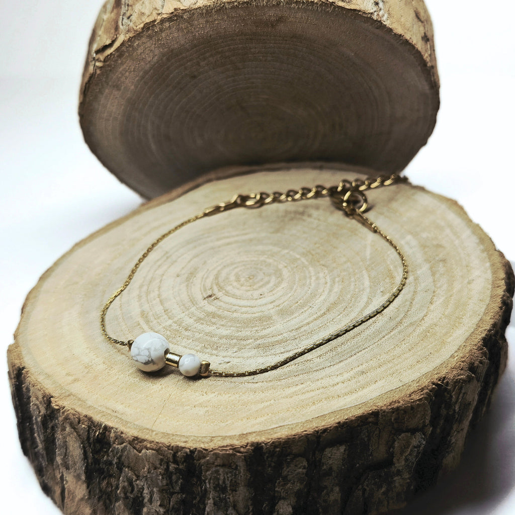 Bracelet fin doré en acier inoxydable avec perles de howlite blanche posé sur fond bois, bijou délicat et apaisant signé Clyvia Bijoux.