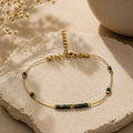 Bracelet fin doré orné de petites perles vert foncé et dorées, posé sur une pierre claire.