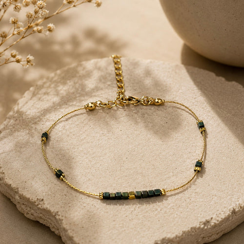Bracelet fin doré orné de petites perles vert foncé et dorées, posé sur une pierre claire.