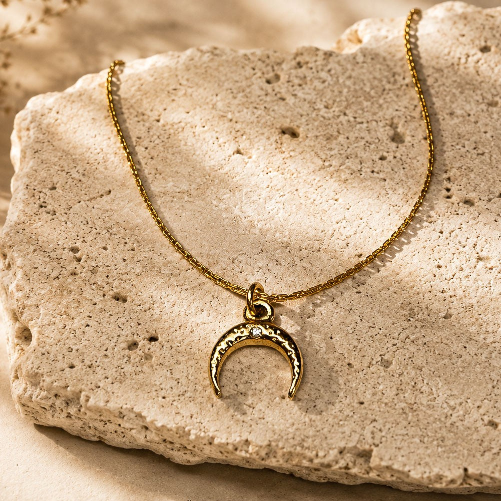 Collier doré à pendentif croissant de lune posé sur une pierre beige texturée.