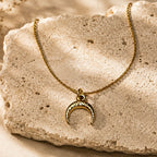Collier doré à pendentif croissant de lune posé sur une pierre beige texturée.