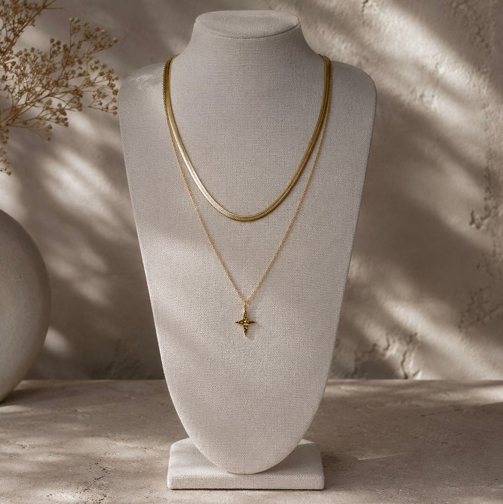 Collier doré à pendentif étoile présenté en superposition avec une chaîne plate sur un buste beige.
