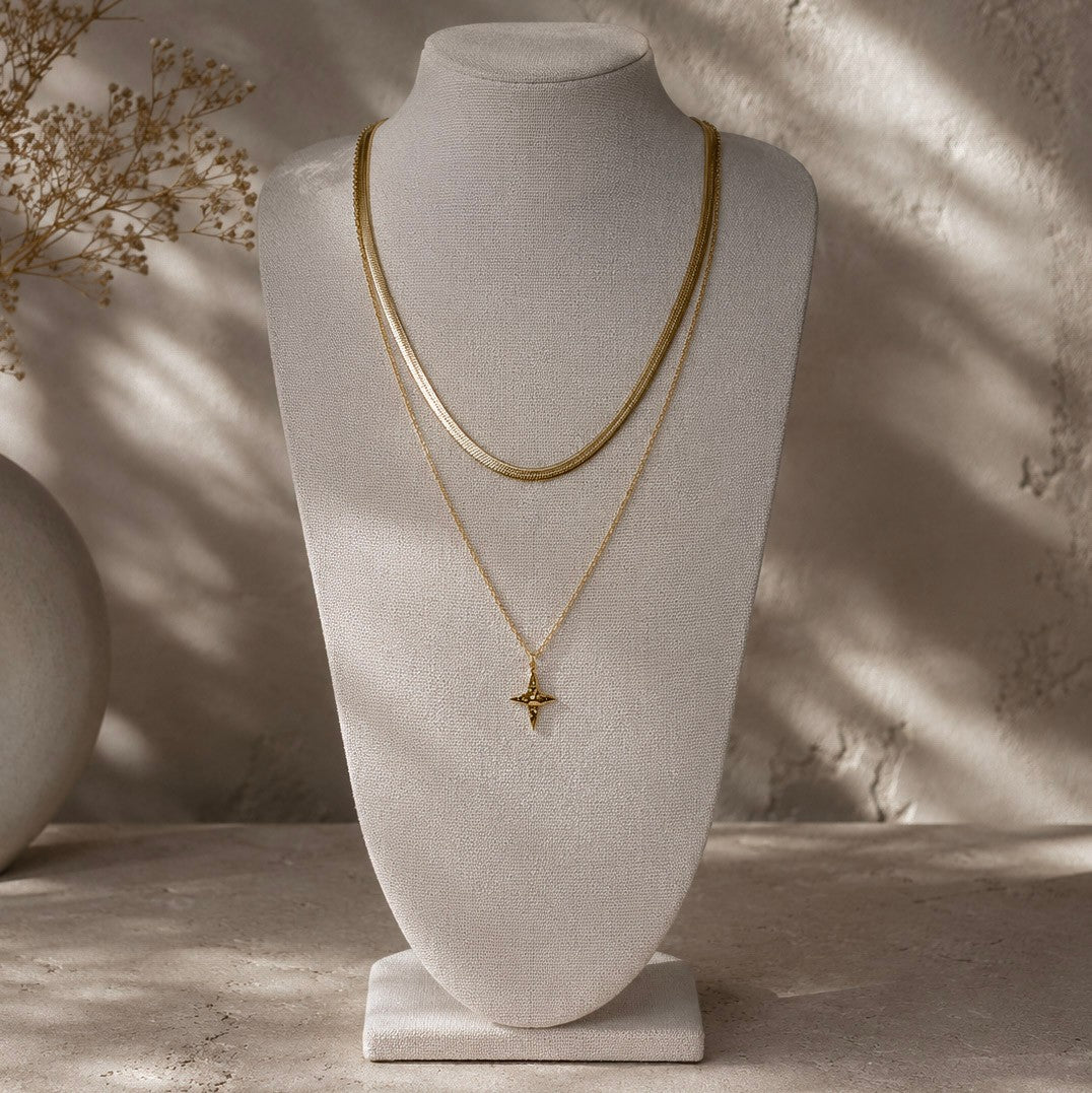 Collier doré à pendentif étoile présenté en superposition avec une chaîne plate sur un buste beige.