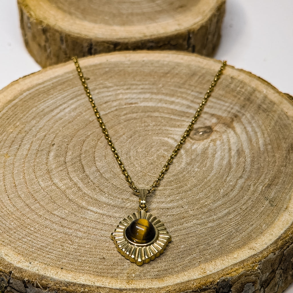 Collier doré en acier inoxydable avec pierre naturelle œil de tigre, pendentif solaire posé sur bois, reflet doré et design intemporel — Clyvia Bijoux.