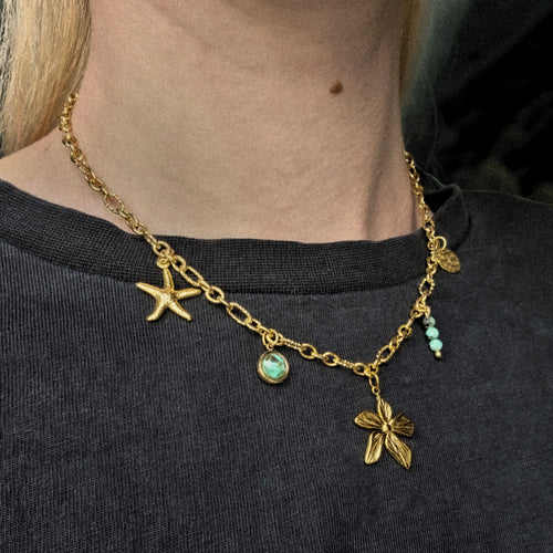 Collier doré en acier inoxydable orné d’une étoile de mer, d’une fleur dorée et de perles en jaspe africaine, porté autour du cou – création artisanale Clyvia Bijoux.