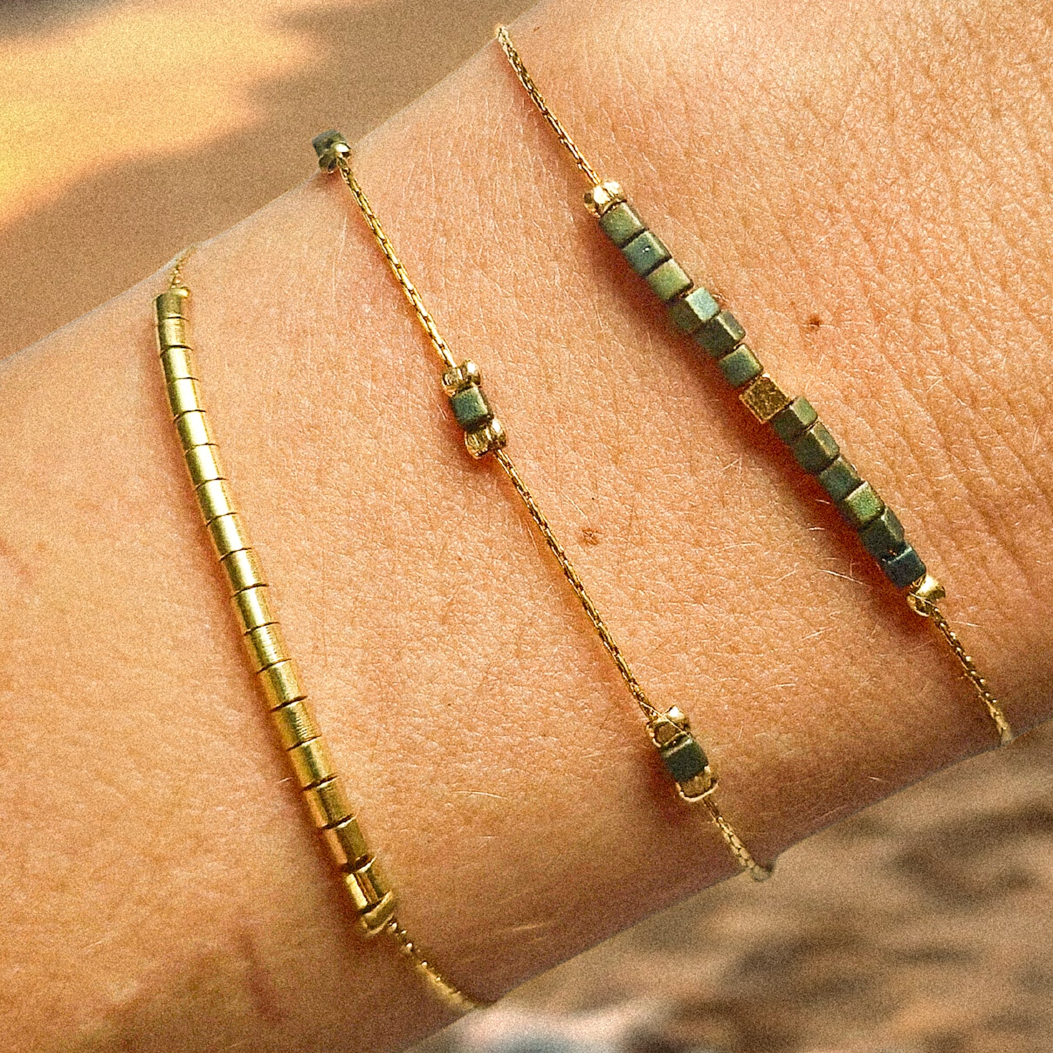 Composition de trois bracelets fins dorés portés au poignet, mélange de perles dorées et de perles en verre irisées vert-bleu, créations élégantes Clyvia Bijoux.